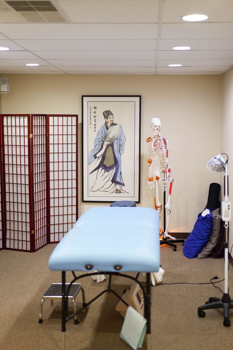 Acupuncture treatment room at Integrative Health Partners Greenville SC — blue treatment table with TDP mineral heat lamp and scalp acupuncture charts on wall