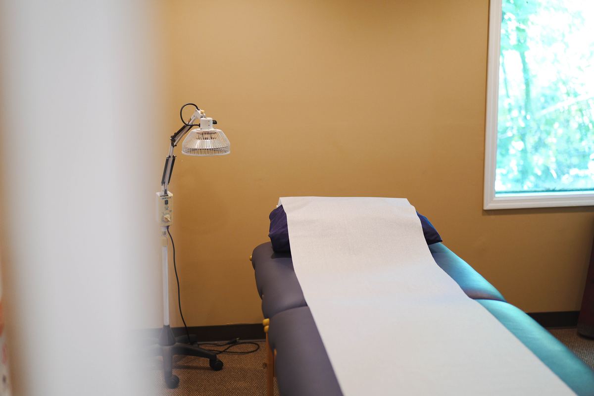 Private acupuncture treatment room at IHP Greenville — TDP mineral lamp beside treatment table with full meridian and acupoint chart on wall, natural light from window